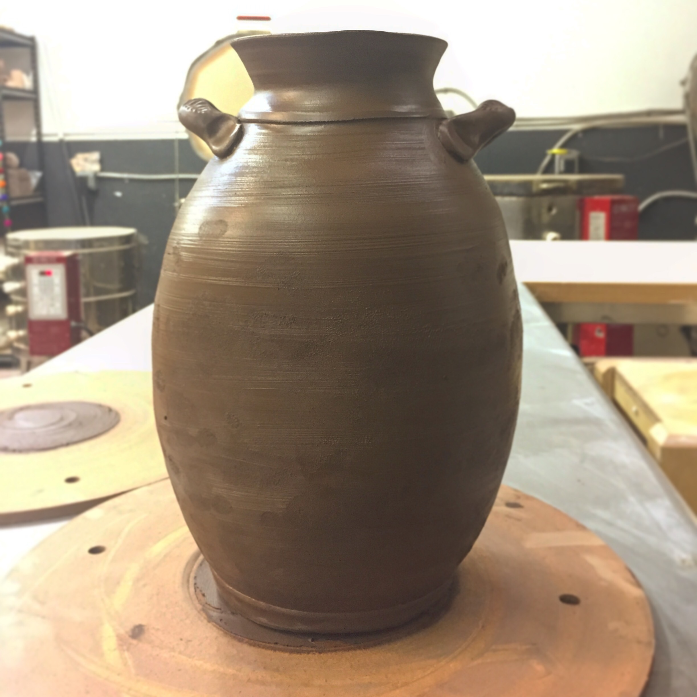 Large clay vase, freshly thrown out of dark brown clay. It's sitting on a throwing bat on a table in a pottery studio.
